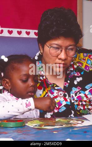Femme hispanique enseignante assise à côté de la fille afro-américaine à l'école pendant que l'enfant fait des aquarelles Banque D'Images