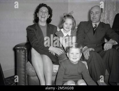 Années 1950, historique, photo de famille, une mère, un père et leurs deux jeunes enfants assis ensemble sur un petit canapé, grands sourires pour leur photo, Angleterre, Royaume-Uni. Banque D'Images