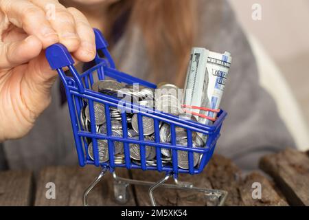 les mains d'une femme âgée tiennent un panier complet d'un supermarché avec des dollars et des pièces sur une table en bois de près Banque D'Images
