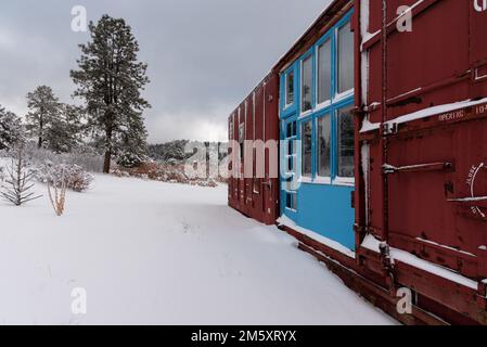 Wagon de train rouge foncé avec de grandes fenêtres et portes encadrées de portes et fenêtres de couleur turquoise dans un paysage enneigé dans le nord du Nouveau-Mexique, États-Unis. Banque D'Images