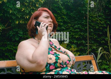 jeune femme latina caucasienne d'origine argentine, plus modèle de taille, cheveux courts de couleur rouge, détendu assis répondant à un appel téléphonique dans le jardin, Banque D'Images