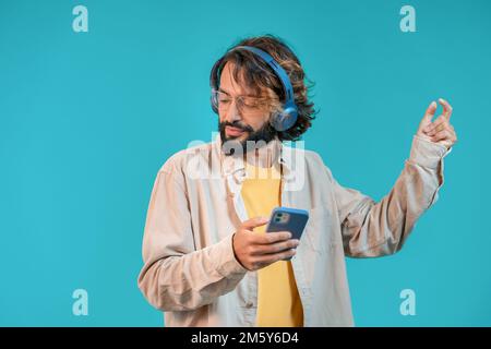 Joyeux homme séduisant dansant sur fond bleu avec un casque et tenant un smartphone Banque D'Images