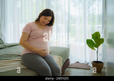 Grossesse, repos des gens et attente concept - asiatique souriant femme enceinte heureuse assis dans le lit et touchant son ventre à la maison. Banque D'Images