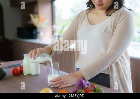 Charmante femme enceinte asiatique tient une bouteille de lait et verse du lait dans un verre. Une femme attirante a besoin de protéines et de calcium pour son bébé et il mak Banque D'Images
