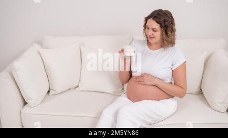 Une femme enceinte s'assoit sur un canapé blanc et boit du lait. Banque D'Images