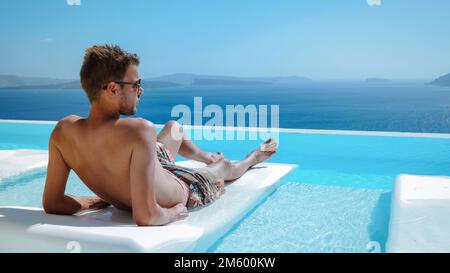 Santorini Grèce Oia, jeunes hommes en short de bain se détendre dans la piscine donnant sur la caldeira de l'île de Santorini Grèce, piscine à débordement, un jeune homme sur des vacances de luxe en Europe Grèce Banque D'Images