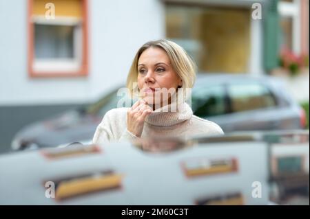 Jolie blonde d'âge moyen, femme sortant d'une voiture et tenant une main sur son col roulé avec un regard réfléchi sur une rue de ville Banque D'Images