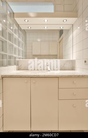 une salle de bains avec des armoires blanches et des plans de travail en marbre sur le meuble-lavabo en face du lavabo est éclairée par des lumières encastrées Banque D'Images
