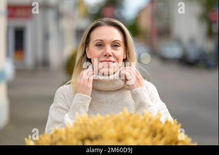 Belle femme blonde attrayante debout souriant derrière des fleurs jaunes avec ses mains sur son turtleneck dans une ville Banque D'Images
