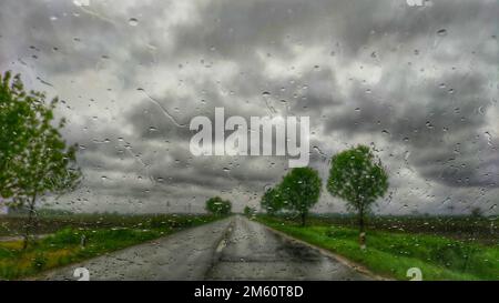 Une route asphaltée entourée d'arbres dans un jour pluvieux vu derrière une voiture wondow avec des gouttes de pluie sur elle Banque D'Images