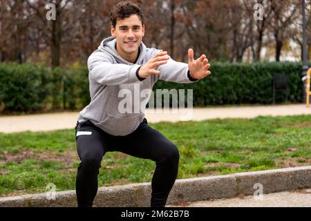 Jeune sportif souriant à capuche grise et leggings noirs faisant des squats dans le parc près des buissons verts tout en regardant l'appareil photo Banque D'Images