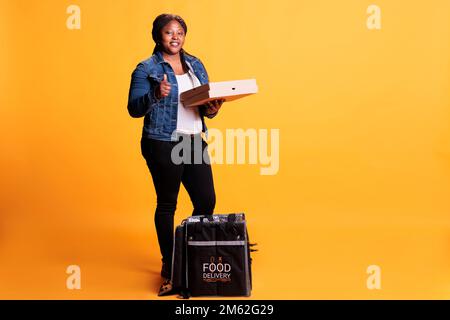 Employé de restaurant réussi tenant la boîte de carton remplie de pizza tout en faisant un geste de pouce vers le haut à l'appareil photo avant de livrer la commande rapide de nourriture au client. Pizzeria coursier transportant un sac à dos thermique à emporter Banque D'Images