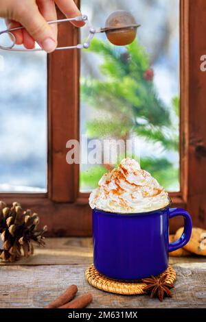poudre de cannelle provenant du tamis volant sur du chocolat chaud avec garniture de crème fouettée dans une tasse en céramique sur une table en bois Banque D'Images