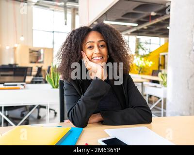 Femme d'affaires afro-américaine positive en tenue officielle assise à une table penchée sur la main pendant la journée de travail regardant la caméra de rêve Banque D'Images
