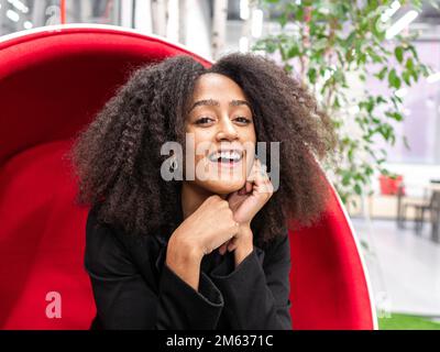 Femme d'affaires noire positive souriant et regardant la caméra tout en se reposant sur une chaise rouge pendant la pause dans un espace de travail moderne Banque D'Images