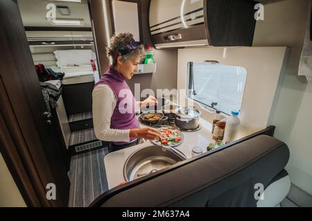 Angle élevé de la femelle dans une tenue décontractée debout dans la cuisine faire une salade délicieuse avec de la laitue avocat et des tomates tout en cuisant le dîner sur la cuisinière pendant Banque D'Images