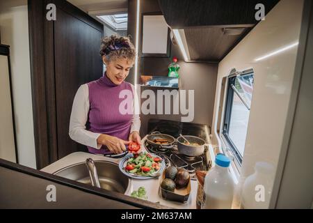 Angle élevé de la femelle dans une tenue décontractée debout dans la cuisine faire une salade délicieuse avec de la laitue avocat et des tomates tout en cuisant le dîner sur la cuisinière pendant Banque D'Images