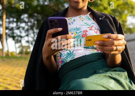 Angle bas de la jeune Afro-américaine dans des boucles d'oreilles de cerceau assis dans la rue près de l'arbre vert et en entrant des références de carte par téléphone cellulaire Banque D'Images