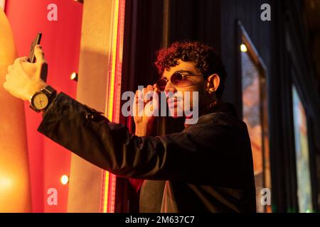 Petit angle de jeune homme dans des vêtements élégants ajuster des lunettes de soleil tout en prenant selfie sur le téléphone portable debout sur la rue de la ville Banque D'Images