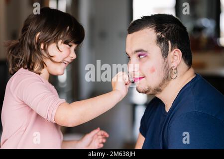 Vue latérale d'une adorable petite fille avec queue de cheval en chandail rose appliquant du maquillage sur le visage du père barbu avec boucle d'oreille en t-shirt noir Banque D'Images