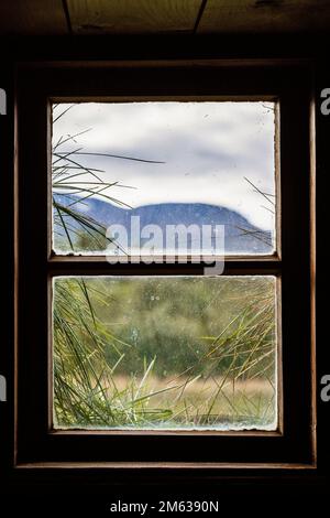 Vue à travers une petite fenêtre avec un cadre en bois sur des plantes vertes et une crête de haute montagne sous un ciel nuageux Banque D'Images