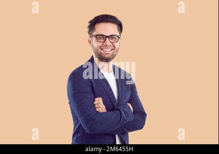 brunet souriant, blouson bleu et lunettes avec main croisée sur la poitrine sur fond beige. Banque D'Images