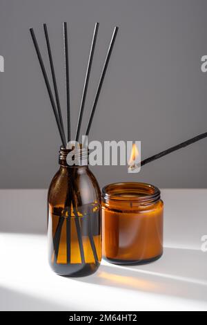 Diffuseur d'arôme avec huile essentielle naturelle en bouteille de verre brun, bougie de cire de soja et de noix de coco avec feu allumé par un match dans un pot ambre sur un blanc gris Banque D'Images