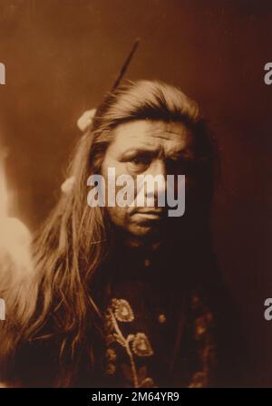 Portrait tête-et-épaules d'un homme de Nespelim, face à face - natif américain, indien - 1904 - Edward S. Curtis, 1868-1952, photographe. Version sépia. Le peuple Nespelem appartient à l'une des douze tribus autochtones confédérées de la réserve indienne de Colville, dans l'est de l'État de Washington. Banque D'Images