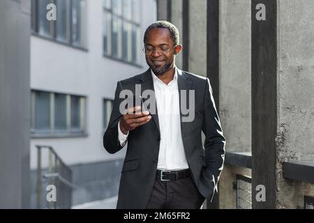 Bon homme d'affaires afro-américain qui a réussi à sortir de l'immeuble de bureaux, directeur tenant un smartphone pour lire des messages et sourire, à l'aide de l'application de communication. Banque D'Images
