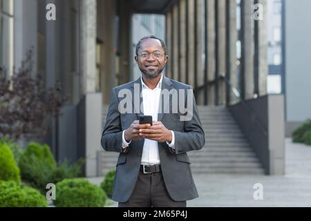 Portrait d'homme d'affaires afro-américain mature, homme d'affaires senior à l'extérieur du bâtiment de bureau tenant le téléphone entre les mains souriant et regardant l'appareil photo. Banque D'Images