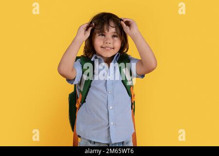 Enfants et problème de vue. Mignon petit garçon d'école avec sac à dos retirer ses lunettes sur le front, fond orange Banque D'Images