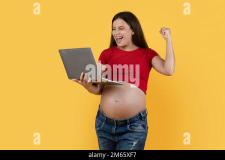 Joyeuse jeune femme enceinte tenant un ordinateur portable et célébrant le succès Banque D'Images
