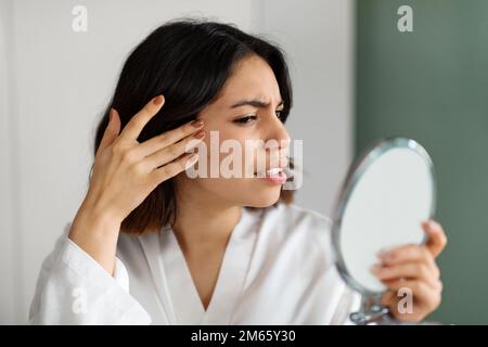 Triste jeune femme souffrant de l'acné, la vérification de la peau dans le miroir Banque D'Images