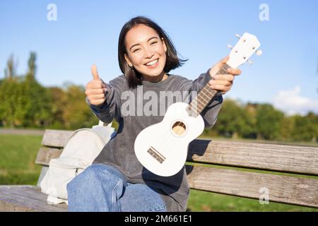 Happy asian girl montre ukulele et pouces vers le haut, montre son nouvel instrument de musique, apprend à jouer dans le parc, assis sur le banc Banque D'Images