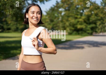 Sourire asiatique de forme physique fille tenant une serviette sur l'épaule, séance d'entraînement dans le parc, la transpiration après les exercices d'entraînement à l'extérieur Banque D'Images