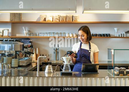 Portrait d'une jeune fille étudiante travaille comme barista, tient la machine à cartes de crédit POS, debout au comptoir avec le terminal et le kit de préparation Banque D'Images