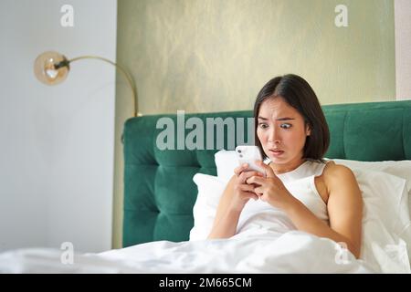 Portrait d'une femme coréenne choquée regardant son téléphone mobile, couché au lit, lisant de mauvaises nouvelles choquantes sur l'application pour smartphone Banque D'Images