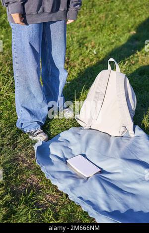 Photo verticale, corps de jeune fille adolescente, debout à côté d'une couverture avec sac à dos et livre, pique-nique à l'extérieur Banque D'Images