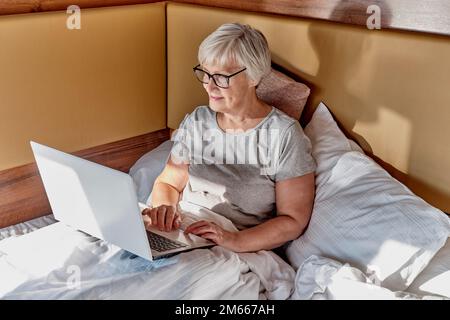 Femme aînée souriante aux cheveux gris courts, utilisant un ordinateur portable dans le lit. À la maison, à l'hôtel, en camping. Travailler en ligne, utiliser des services en ligne, en acheter Banque D'Images