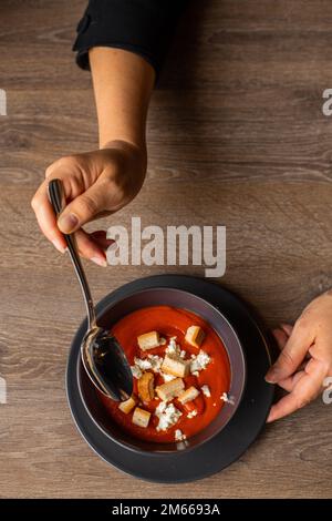 Photo verticale de la femme coupée les mains remuent la soupe à la crème de tomate rouge avec des toasts de croûton de soupe sur une assiette noire sur une table en bois Banque D'Images