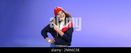 Photo en studio d'une charmante femme enthousiaste aux cheveux bouclés, invitant à jouer dans la neige avec elle pointant vers le coin supérieur droit, tête inclinable joyeusement et Banque D'Images