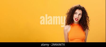 Élégante femme aux cheveux chauds et foncés avec boucles et rouge à lèvres dans un haut court tendance regardant dans le coin supérieur gauche intrigué et curieux de clenching dents Banque D'Images