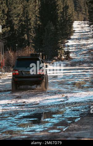 Vue arrière en angle du tout-terrain Overland Land Cruiser SUV croisant l'eau sur un sentier enneigé dans la forêt Banque D'Images