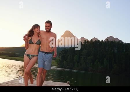 L'amour et le rire au lac. un jeune couple affectueux en maillots de bain marchant sur un quai au coucher du soleil. Banque D'Images