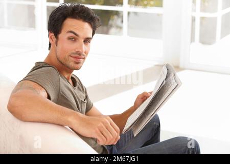 Rester informé des dernières actualités. Un beau jeune homme assis sur le canapé à la maison pour lire un journal. Banque D'Images