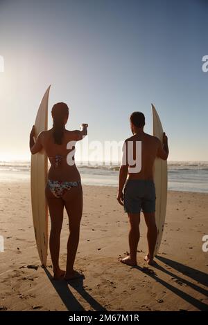 Shes a trouvé la meilleure pause. Vue arrière d'un jeune couple de surfeurs regardant la plage et les vagues lointaines. Banque D'Images