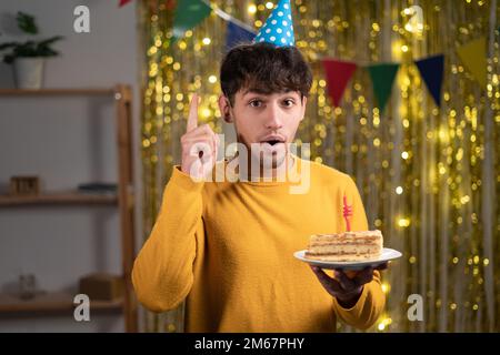 Jeune arabe avec cheveux bouclés portant un chapeau d'anniversaire tenant un gâteau à l'intérieur de la maison surpris par une idée ou une question pointant du doigt avec le visage heureux Banque D'Images