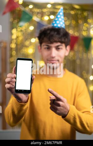 Concept de maquette. Beau homme arabe confiant en chandail jaune et chapeau de fête montrant un smartphone avec écran vide, pointe le doigt sur l'appareil vierge Banque D'Images