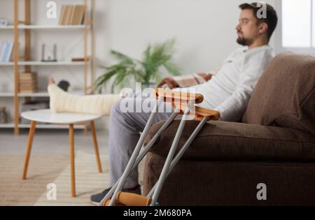 Jeune homme avec une jambe blessée assis à la maison sur un canapé en réadaptation avec ses béquilles Banque D'Images