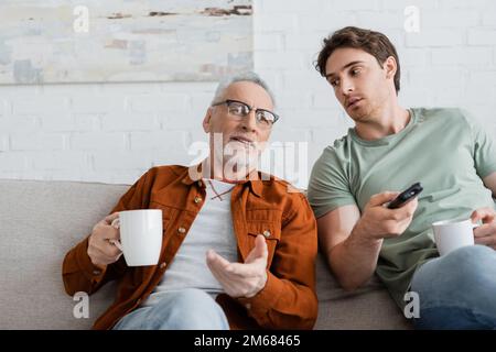 homme mûr avec tasse de thé pointant avec la main près de son en cliquant sur les chaînes tv sur la télécommande, image de stock Banque D'Images